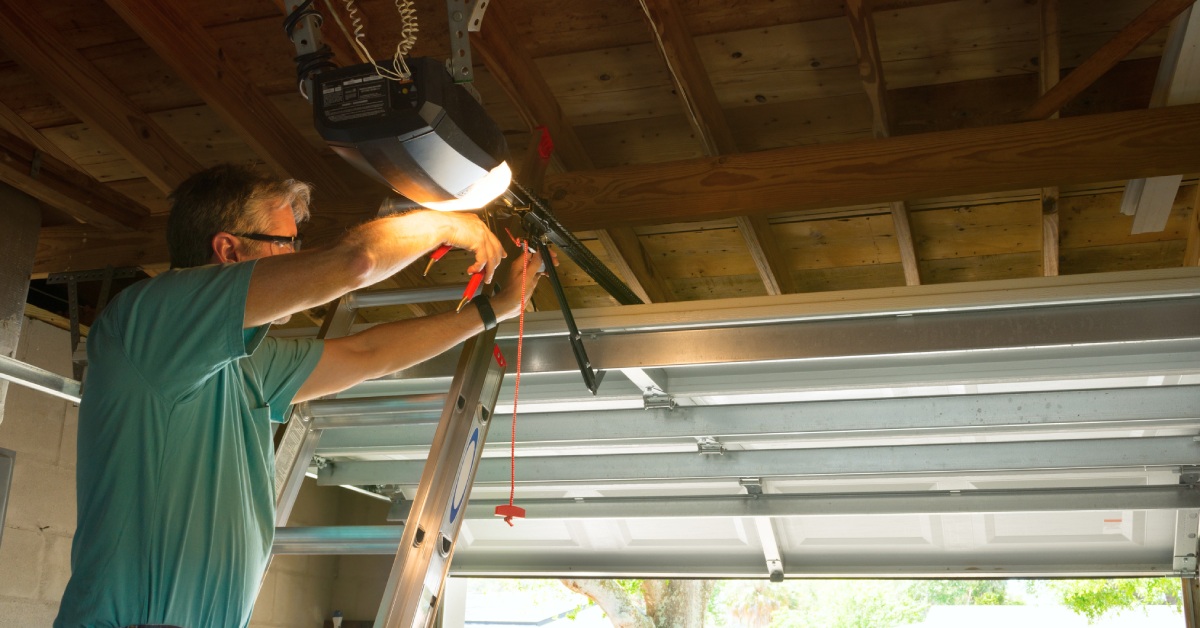 A person wearing safety glasses standing on a ladder to adjust a garage door opener with a hand tool. The garage door is open.