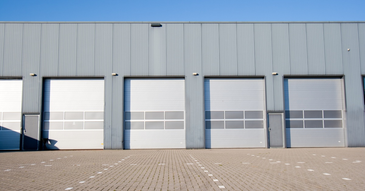 A warehouse with five single overhead garage doors. Lanes for the doors are white dotted lines on the pavement.